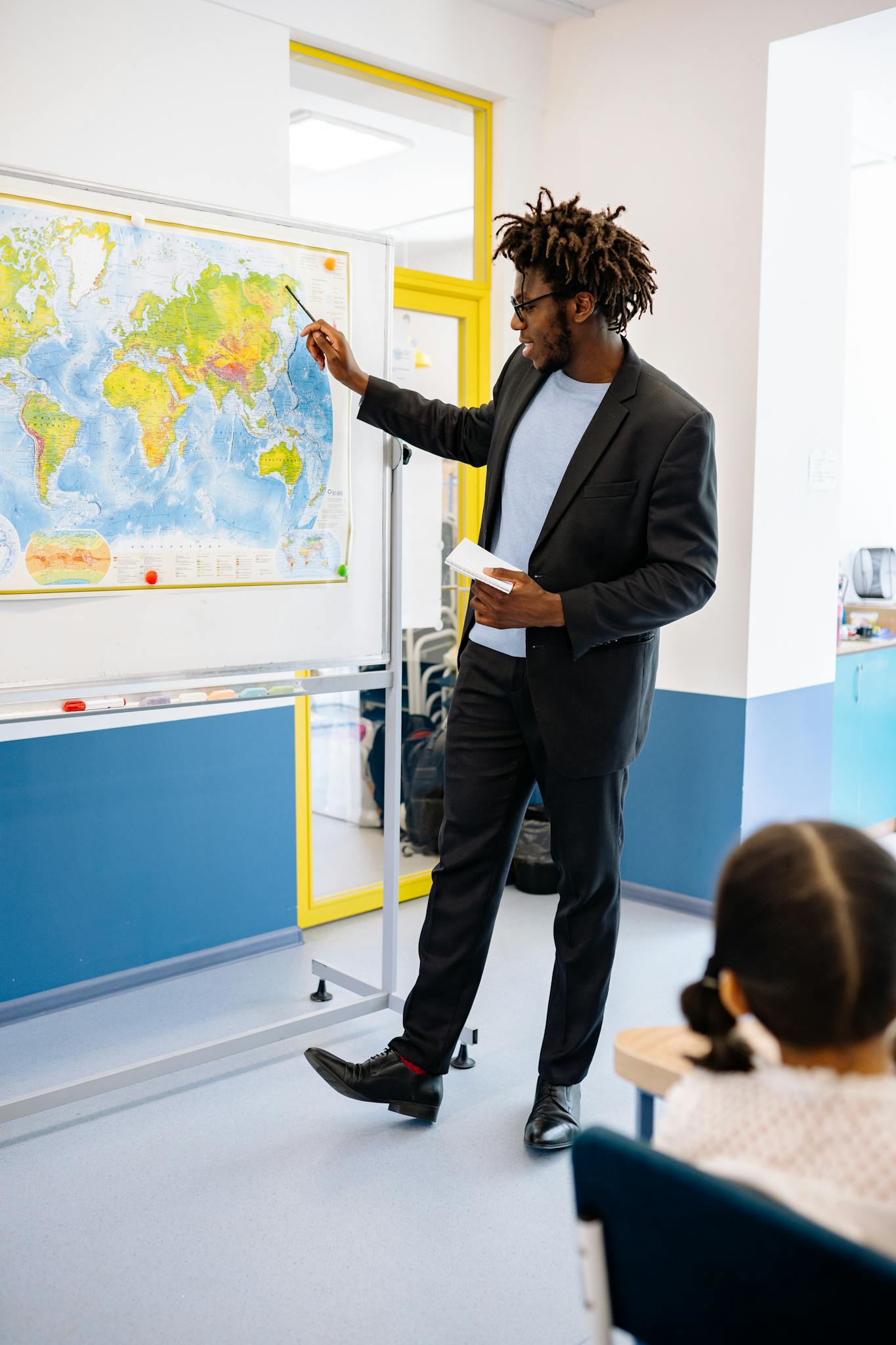 A male teacher explains world geography using a map to students in a classroom.